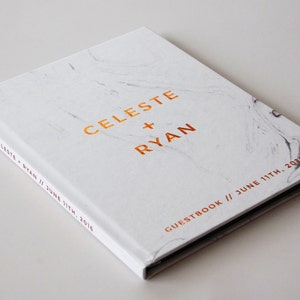 May include: A white marble-patterned guestbook with the names "CELESTE + RYAN" in gold lettering. The date "JUNE 11TH, 2016" is also displayed. The book's spine reads "CELESTE + RYAN // JUNE 11TH, 2016".