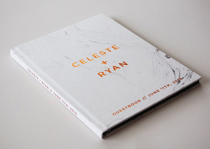 May include: A white marble-patterned guestbook with the names "CELESTE + RYAN" in gold lettering. The date "JUNE 11TH, 2016" is also displayed. The book's spine reads "CELESTE + RYAN // JUNE 11TH, 2016".