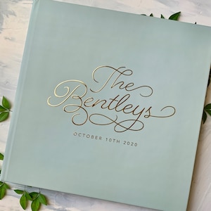May include: A light blue, square photo album with gold foil lettering that reads "The Bentleys" and "October 10th 2020". The album is surrounded by green leaves.