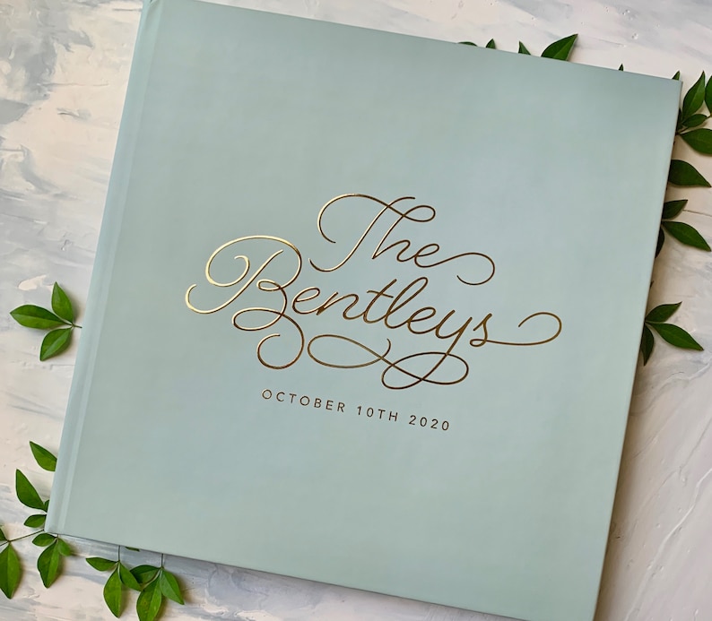 May include: A light blue, square photo album with gold foil lettering that reads "The Bentleys" and "October 10th 2020". The album is surrounded by green leaves.