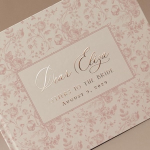 K&ouml;nnte beinhalten: Ein Hardcover-Buch mit einem floralen Muster in zartem Rosa und Creme. Auf dem Einband steht in eleganter Schrift "Dear Eliza", darunter "Letters to the Bride" und das Datum 9. August 2029. Das Buchdesign ist romantisch und vintage-inspiriert.
