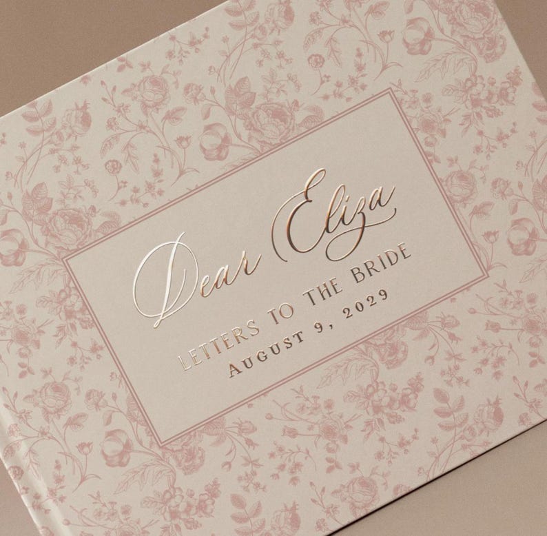 K&ouml;nnte beinhalten: Ein Hardcover-Buch mit einem floralen Muster in zartem Rosa und Creme. Auf dem Einband steht in eleganter Schrift "Dear Eliza", darunter "Letters to the Bride" und das Datum 9. August 2029. Das Buchdesign ist romantisch und vintage-inspiriert.