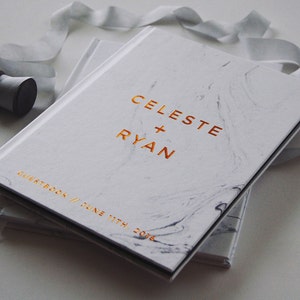 May include: A white marble-patterned guestbook with gold lettering that reads "CELESTE + RYAN" and "GUESTBOOK // JUNE 11TH, 2016". A gray ribbon and spool of thread are next to the book. The book is closed.