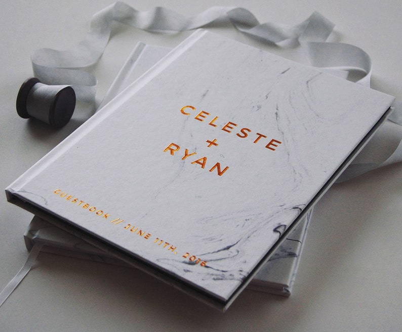 May include: A white marble-patterned guestbook with gold lettering that reads "CELESTE + RYAN" and "GUESTBOOK // JUNE 11TH, 2016". A gray ribbon and spool of thread are next to the book. The book is closed.