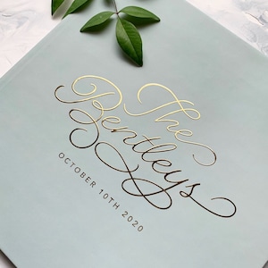 May include: A light blue book cover with gold script text that reads "The Bentleys" and the date "October 10th 2020". A sprig of green leaves is in the upper corner.