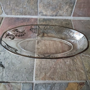 May include: Clear oval glass serving dish with a silver rim and floral design. The dish features a delicate pattern of flowers and leaves, adding a touch of elegance. This decorative piece is suitable for serving appetizers or desserts.