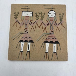 May include: A square, tan-colored tile featuring a sand painting of two stylized figures. The figures have geometric heads, patterned bodies, and are flanked by green plant-like shapes. The artwork is in shades of brown, red, black, and white.