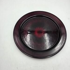 May include: A round, deep red glass plate featuring a smooth, reflective center with a radiating starburst pattern. The wide rim is textured with a fine, cross-hatched grid design that transitions into a darker, translucent burgundy hue.