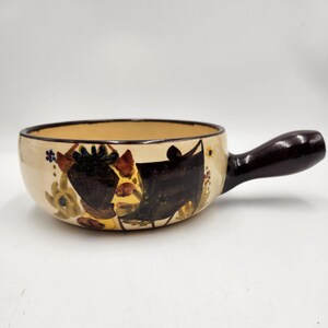 May include: Cream-colored ceramic soup bowl with a dark brown handle. The bowl features a hand-painted floral design in shades of brown, yellow, and blue. The interior of the bowl is a light tan color.