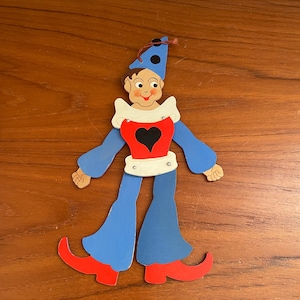 May include: A vintage, whimsical cutout of a clown figure. The clown has a blue hat with black dots, a red heart on the chest, and blue pants with red shoes. The figure is set against a wooden background.