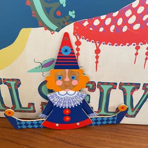 May include: A colorful wooden clown decoration with a red and blue striped hat, orange hair, and a blue outfit. The clown has a white collar and is set against a backdrop with a whimsical design. The clown's arms are curved and blue with a diamond pattern.