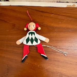 May include: A wooden toy doll ornament with a red and white checkered dress, red pants, and black shoes. The doll has a painted face with yellow hair and red cheeks.