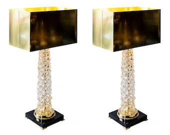 Puede incluir: Un par de lámparas de mesa doradas con bases de cristal transparente y bases cuadradas negras. Las lámparas tienen pantallas rectangulares.