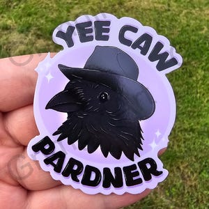 May include: A purple and black sticker featuring a cartoon crow wearing a cowboy hat. The sticker reads "Yee Caw Pardner" in a black, bold font.