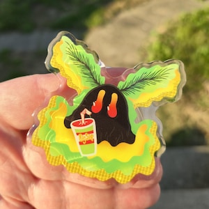 May include: A clear, stylized pin featuring a black blob character with red feet and a drink. The character is surrounded by green and yellow foliage. The drink cup has the word "Magic" on it.