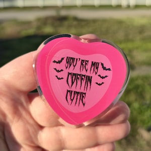 May include: A pink heart-shaped acrylic item with the text "YOU'RE MY COFFIN CUTIE" in black gothic lettering, and small bat illustrations. The item is held in a person's hand.