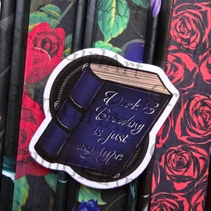 Puede incluir: Una pegatina con un diseño de libro azul oscuro. El libro tiene una cadena dorada y el texto "Dark & Brooding is just my type".
