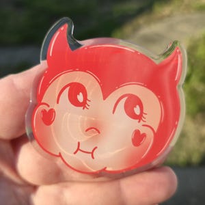 May include: A clear acrylic pin featuring a cartoon devil face with red horns, cheeks, and a winking eye. The pin has a heart-shaped blush on each cheek and a smiling mouth. The background is blurred green.