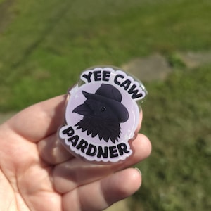 May include: A circular sticker featuring a cartoon crow in a cowboy hat. The text "Yee Caw Pardner" is written around the crow. The crow is black with a purple background. The sticker is clear.
