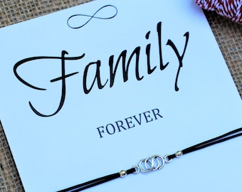 May include: A white card with the word "Family" in elegant script, with "FOREVER" below. A black string bracelet with silver interlocking circles and gold beads is attached. The card includes the text "The love of a Family is life's greatest blessing!" and "May your wish come true!"