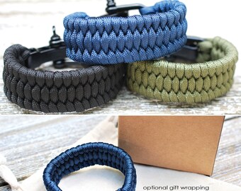 Puede incluir: Tres pulseras de paracord en azul, negro y verde con cierres metálicos negros. Las pulseras están apiladas una encima de la otra. La imagen también muestra una caja marrón y una bolsa blanca, que son opciones de envoltorio de regalo opcionales.