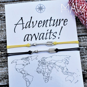 May include: Two friendship bracelets with arrow charms. The bracelets are black and yellow and the charms are silver. The bracelets are on a white card with the text "Adventure awaits!" and a world map.