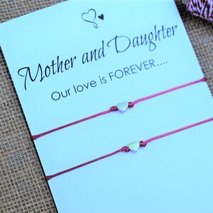 May include: Two matching pink string bracelets with silver heart charms are displayed on a card that reads "Mother and Daughter, Our love is FOREVER...." The background is a light brown burlap.