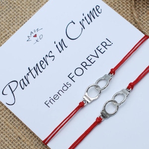 May include: Two red string bracelets with silver handcuff charms. The bracelets are on a white card with the text "Partners in Crime Friends Forever!"
