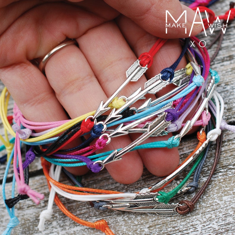 May include: A collection of colorful string bracelets with silver arrow charms. The bracelets are arranged in a hand, with the charms facing the viewer. The bracelets are made of thin string and have a simple design. The text "MAKE A WISH XO" is visible on the top of the image.