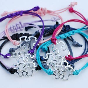 May include: A set of colorful friendship bracelets with a silver puzzle piece charm. The bracelets are made of different colors of cord, including purple, pink, lavender, teal, navy, and red.