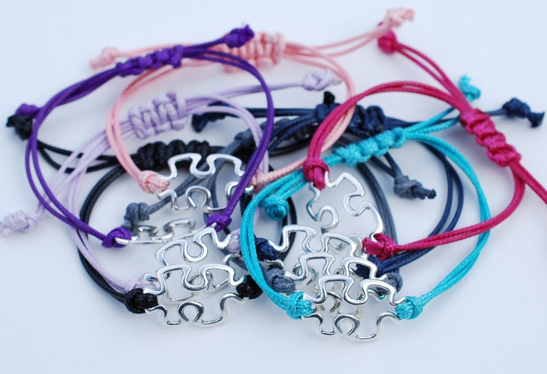 May include: A set of colorful friendship bracelets with a silver puzzle piece charm. The bracelets are made of different colors of cord, including purple, pink, lavender, teal, navy, and red.