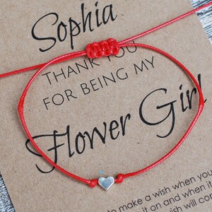 May include: Red string bracelet with a silver heart charm. The bracelet is on a card that says "Sophia, Thank you for being my Flower Girl!"