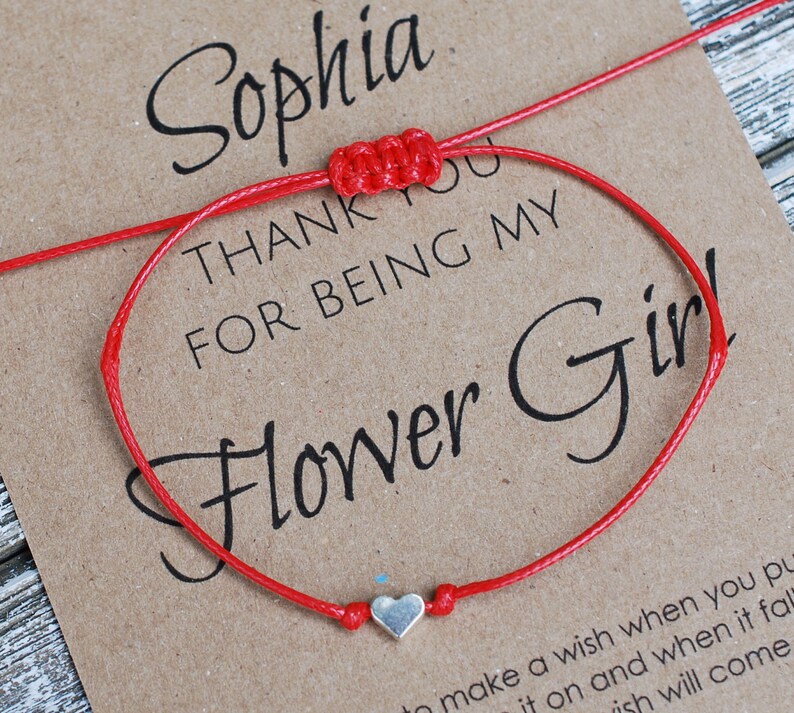 May include: Red string bracelet with a silver heart charm. The bracelet is on a card that says "Sophia, Thank you for being my Flower Girl!"
