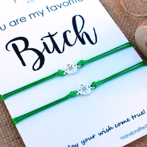 May include: Two green friendship bracelets with silver compass charms. The bracelets are on a white card with the text "You are my favorite Bitch" and "May your wish come true! Handcrafted with love in USA."