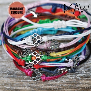 May include: A stack of colorful bracelets with macrame closures and silver metal charms. The bracelets are made of thin cord and are arranged in a rainbow pattern.