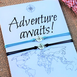 May include: Two matching bracelets with compass charms, one light blue and one black, are displayed on a card with a world map and the words "Adventure awaits!". The bracelets are tied with adjustable knots.