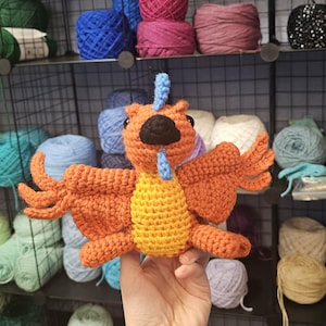 May include: A handmade orange, yellow, and blue crochet bird with outstretched wings. The bird is held in front of a shelf filled with various colors of yarn. The yarn is in shades of blue, purple, pink, green, and white.