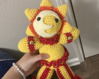 May include: A crocheted sun-shaped plush toy with a yellow body, red and orange accents, and a white crescent moon on its face. The toy is wearing red and yellow striped trousers with ruffled edges and has bells on its feet.