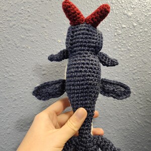Könnte beinhalten: Eine handgefertigte Häkelfigur in Marineblau mit roten Hörnern. Das Plüschtier hat kleine Flügel und einen langen Schwanz mit dreizehigen Füßen. Das Spielzeug wird in einer Hand gehalten.