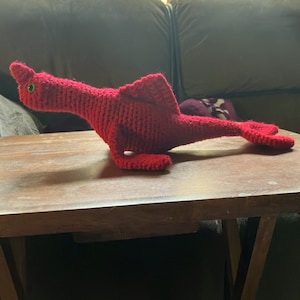 May include: A red crocheted seahorse with a green eye, lying on a wooden table.