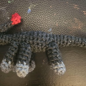 Könnte beinhalten: Handgefertigte Häkeltierfigur in Grautönen mit weißen und schwarzen Augen und einem kleinen roten Akzent. Das Spielzeug hat vier Beine und einen langen Schwanz. Die Kreatur liegt auf einer dunklen Oberfläche.