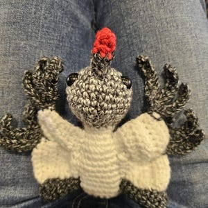 Puede incluir: Un pájaro de ganchillo hecho a mano con una cresta roja, cuerpo gris y blanco y ojos negros. Las alas y las plumas de la cola son grises y blancas. El pájaro está sobre una superficie de mezclilla azul.