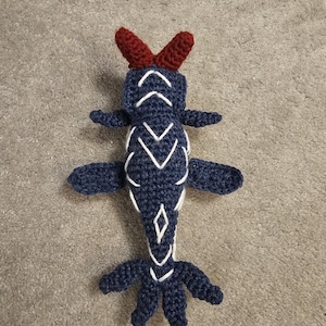 May include: A dark blue crocheted creature with two dark red horn-like protrusions on its head. The body features white embroidered chevron and diamond patterns, four side fins, and a multi-pointed tail. The texture is yarn-based and handmade.