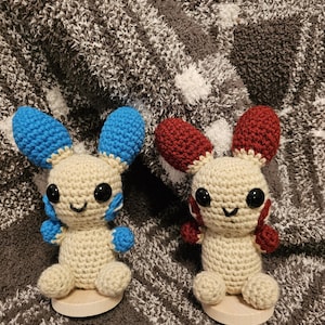 May include: Two handmade crochet bunny dolls. One bunny has blue ears and accents, the other has red ears and accents. Both bunnies have beige bodies, black eyes, and are sitting on small wooden bases.