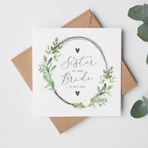 May include: A square greeting card with the text "Sister of the Bride" inside a floral wreath. The card includes the date "18 May 2024" and is accompanied by a brown envelope. The wreath features green leaves, white flowers, and two black hearts.