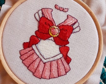Op de afbeelding: Een roze en rode kruissteekborduursel van een sailor scout-outfit met een grote rode strik en een gouden ster broche.