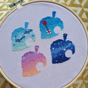 May include: Four embroidered Animal Crossing leaf designs on a white fabric stretched over a wooden embroidery hoop. The designs are a blue leaf with a red balloon, a blue leaf with stars, a blue and green leaf with clouds, and a pink and orange leaf with a sunset.