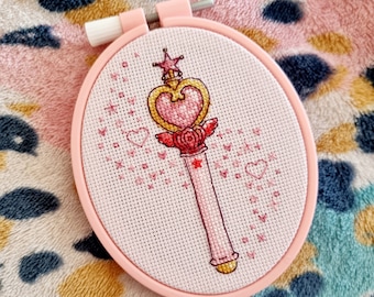 Op de afbeelding: Een roze en gouden geborduurde hoepelkunstwerk met een roze hartvormige toverstaf met een ster erop. De toverstaf is omringd door kleine roze hartjes.