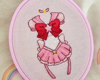 Op de afbeelding: Een geborduurd Sailor Scout-kostuum in roze en rood met een gele ster op de rok en een gele ster op de strik. De borduurwerk is in een hoepel.