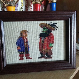 Könnte beinhalten: Ein gerahmtes Kreuzstichbild mit zwei pixelartigen Figuren. Die Figur links ist ein Mann mit braunen Haaren und einer blauen Jacke. Die Figur rechts ist ein grünhäutiger Mann mit einer roten Jacke und einem großen Hut.
